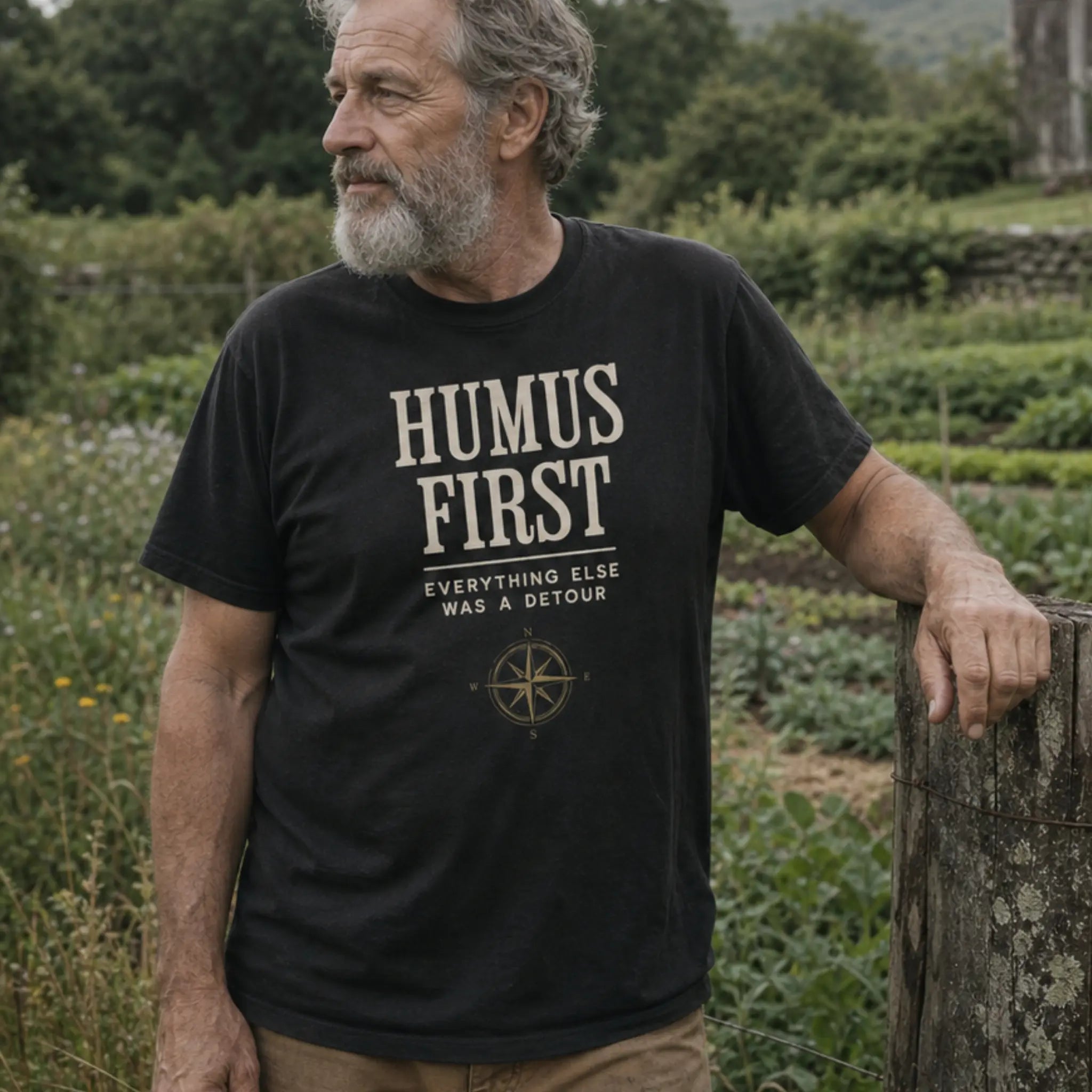 Humus First organic cotton t-shirt worn by man leaning on a fence post on a regenerative farm with vegetable beds