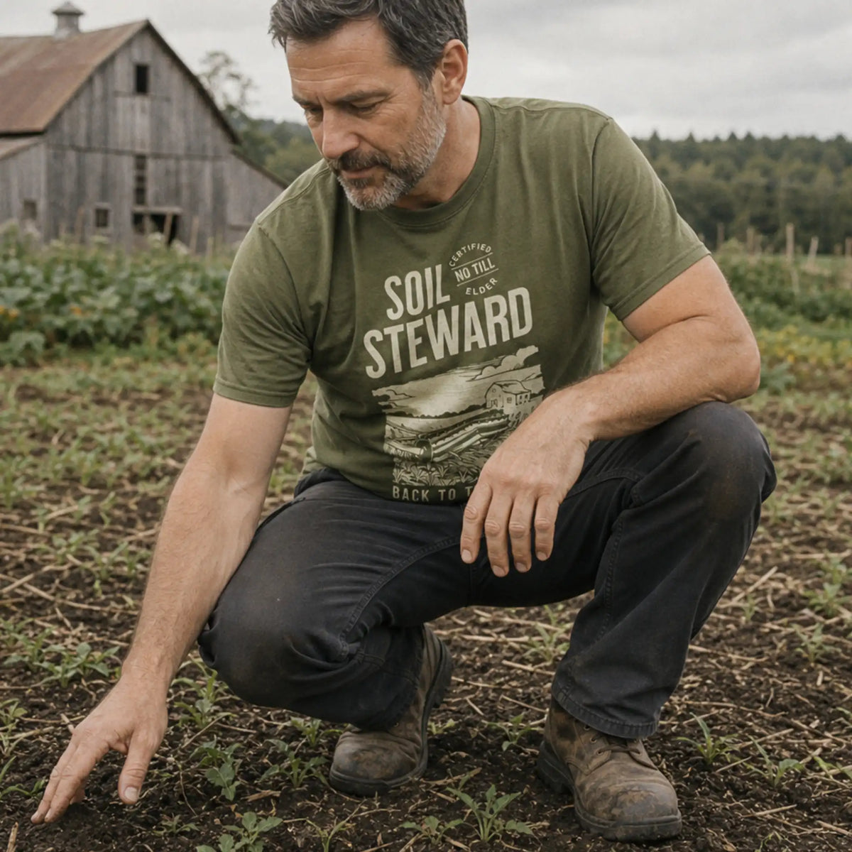 No Till Elder, Soil Steward organic cotton t-shirt worn by man crouching in a field with young seedlings and a barn in the background