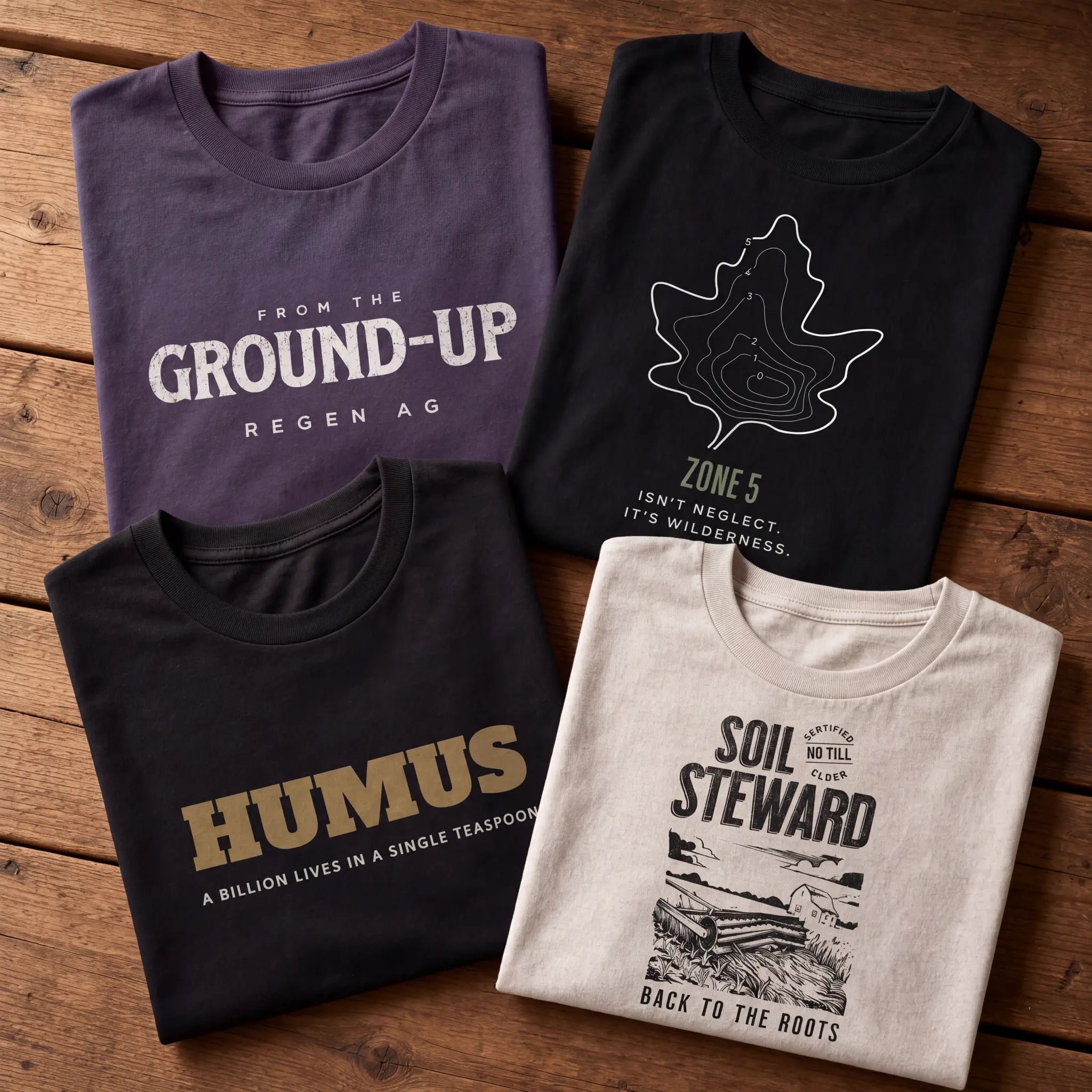 Four folded Emogine organic cotton t-shirts on a wooden table showing Regenerative Agriculture, Zone 5 Wilderness, Humus Microbiology and No Till Elder designs — GOTS-certified regenerative agriculture apparel