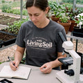 Woman wearing a charcoal Living Soil T-shirt while taking notes beside a microscope in a greenhouse, with plants and growing beds in the background.
